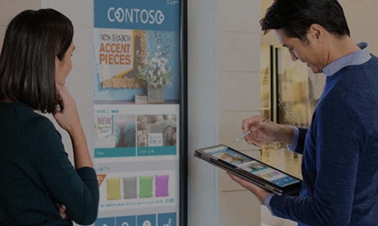 a man holding a tablet and a woman standing in front of a digital screen showing store items (cushions)
