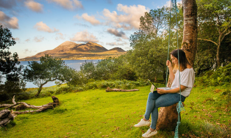 Woman in white t-shirt and jean on a swing working on a laptop with a lake an mountain in the background