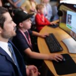 A person in a suit sits beside a child using a computer, with several other people working at computers in the background.