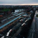 Flygbild över ett stadsområde med en större knutpunkt för bussar och tåg, intill en bred väg. Byggnader, grönområden och vatten syns i bakgrunden.