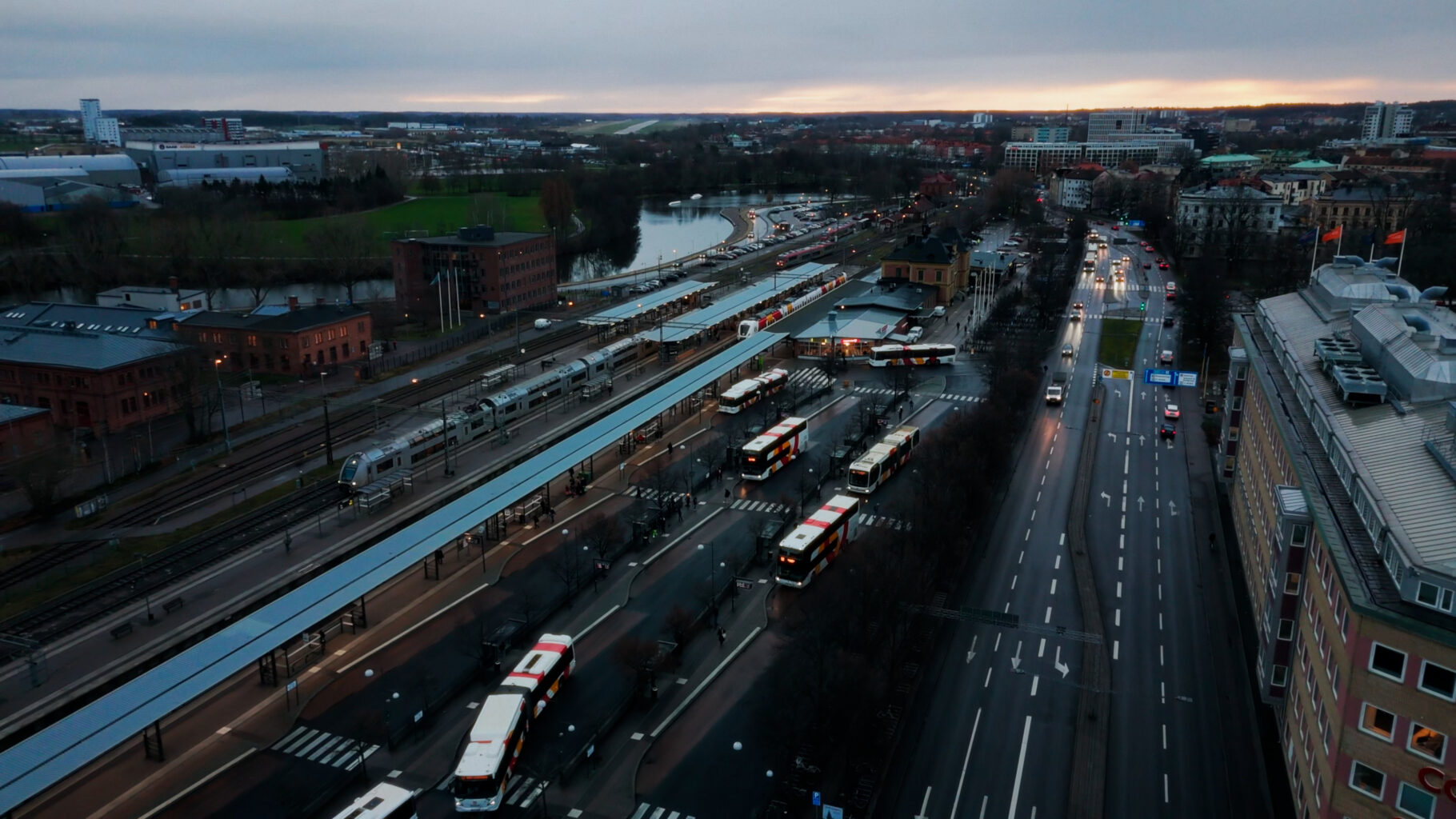 Flygbild över ett stadsområde med en större knutpunkt för bussar och tåg, intill en bred väg. Byggnader, grönområden och vatten syns i bakgrunden.