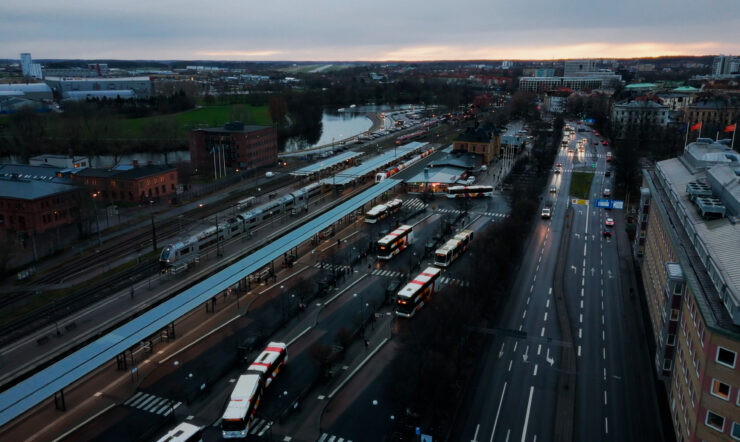 Flygbild över ett stadsområde med en större knutpunkt för bussar och tåg, intill en bred väg. Byggnader, grönområden och vatten syns i bakgrunden.