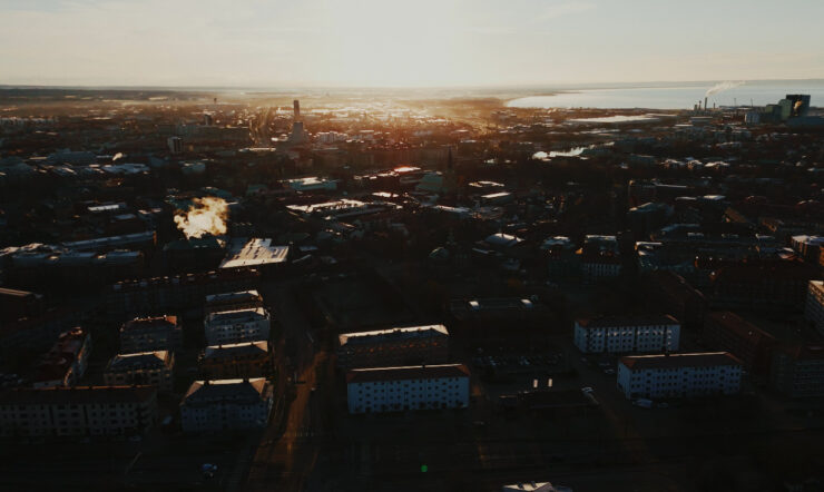 Flygbild över en stad i kvällsljus, med låga byggnader i förgrunden och havet vid horisonten. Solen står lågt över staden.