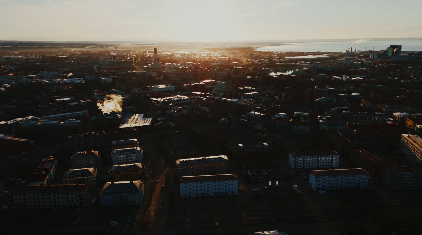 Flygbild över en stad i kvällsljus, med låga byggnader i förgrunden och havet vid horisonten. Solen står lågt över staden.