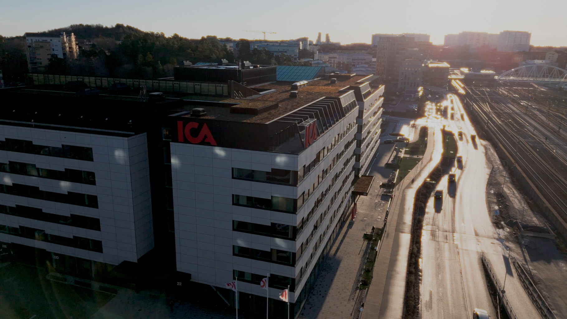 Aerial view of an ICA-branded office building beside a road and multiple railway tracks, with the city extending into the background in warm evening light.