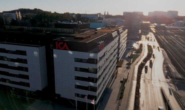 Aerial view of an ICA-branded office building beside a road and multiple railway tracks, with the city extending into the background in warm evening light.