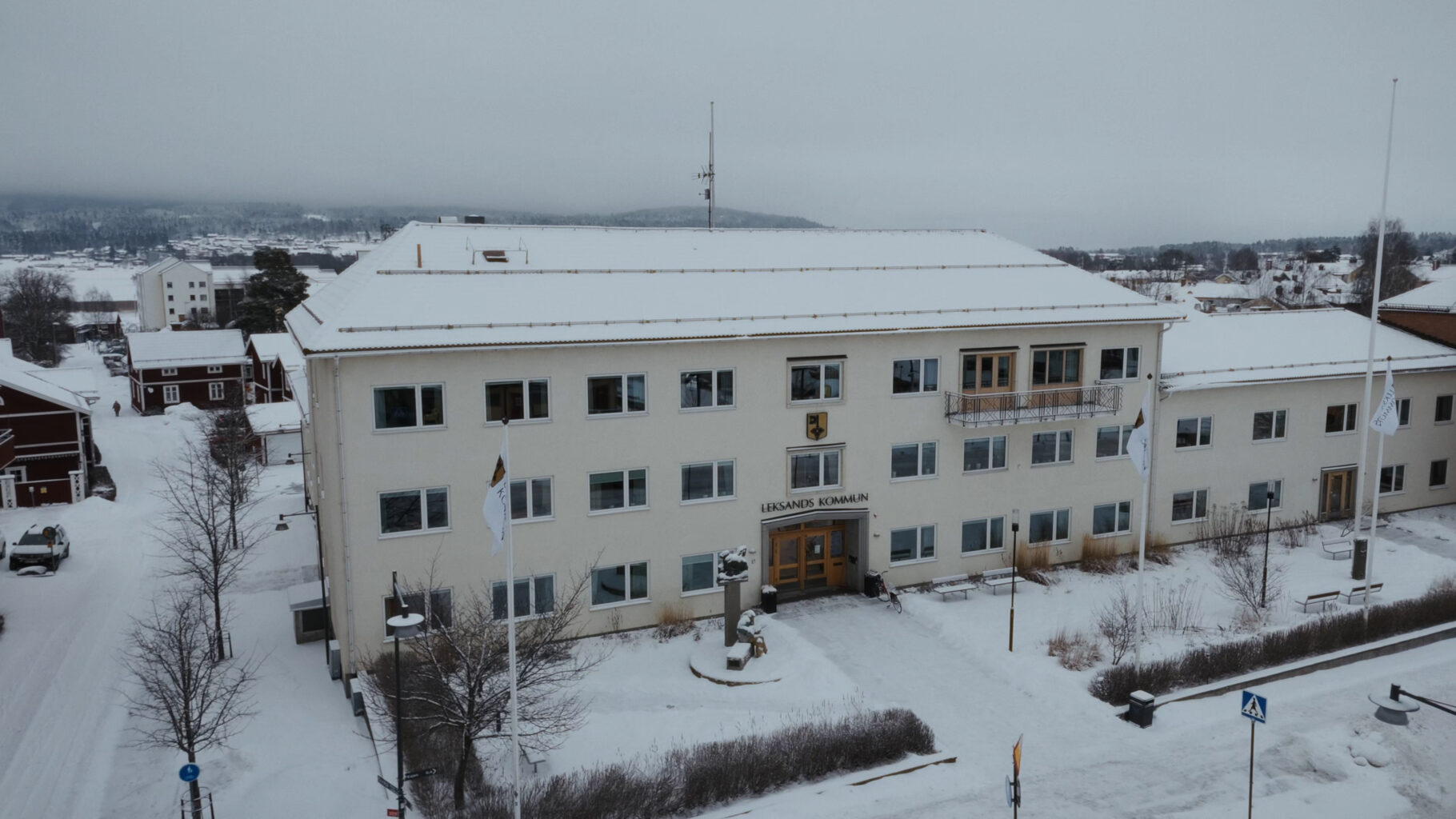 Flygbild över en stor vit byggnad i ett snötäckt landskap, med flaggstänger och entré framför byggnaden samt mindre byggnader och träd i bakgrunden.