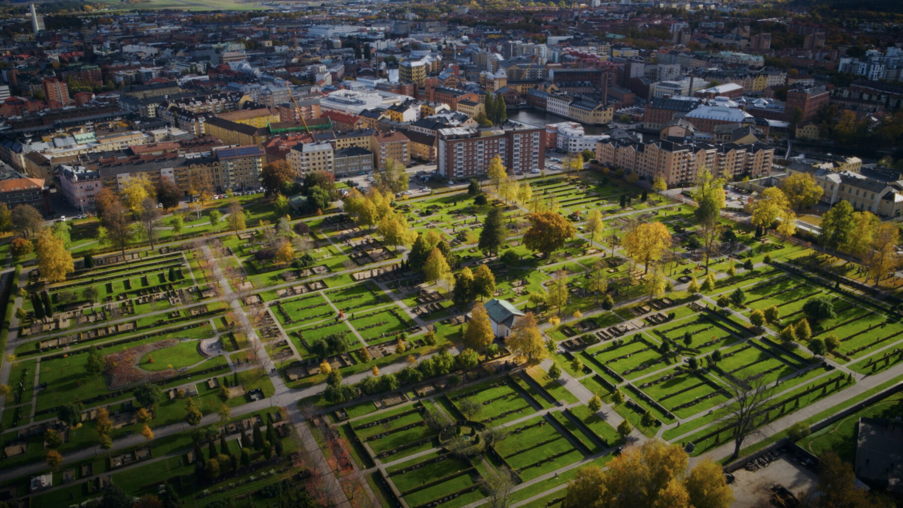 Flygbild över en stor kyrkogård med gröna gräsmattor, gångvägar, gravplatser och träd. Tät stadsbebyggelse syns i bakgrunden.
