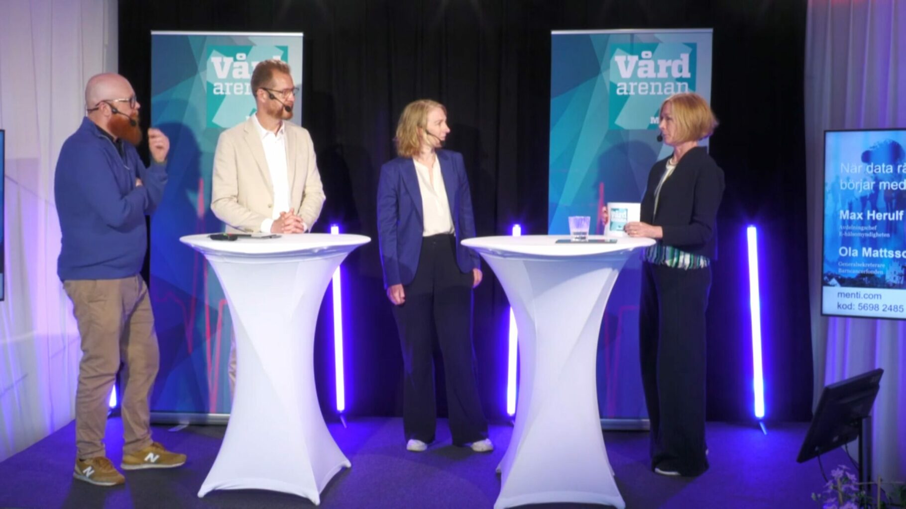 Fyra personer står på en scen och samtalar vid två höga, vita ståbord. I bakgrunden syns två skyltar med texten ”Vårdarenan”, svarta draperier och vertikala ljuslister i blått och lila. Till höger syns även en skärm med text och namn.
